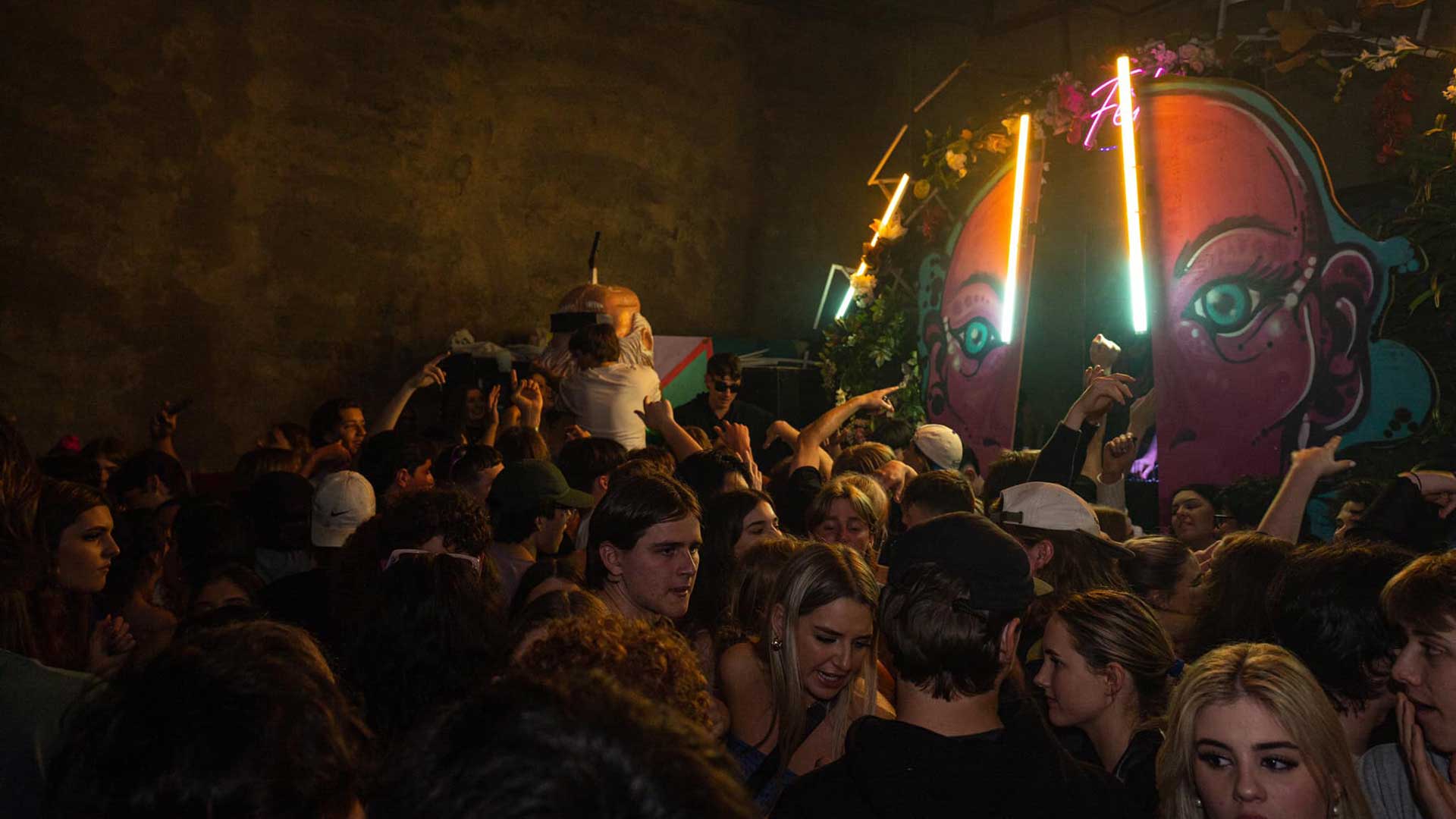 an electronic music event in a warehouse packed full of ravers around a large stage backdrop of a cartoon character adorned with fluorescent lights and flowers
