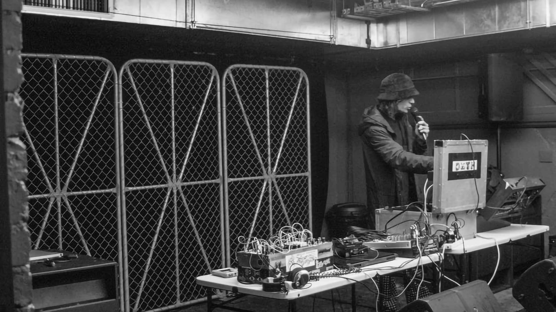 a black an white image of an electronic male artist performing on modular synths holding a microphone  