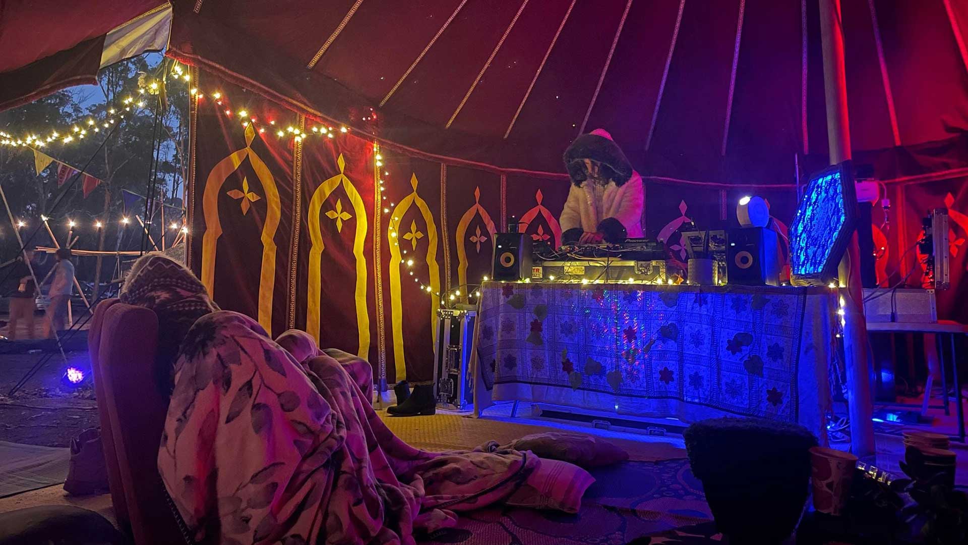 an ambient electronic artist wearing a white furry robe performing on DJ equipment in a large circus tent decorated with ambient lighting and music equipment. 
