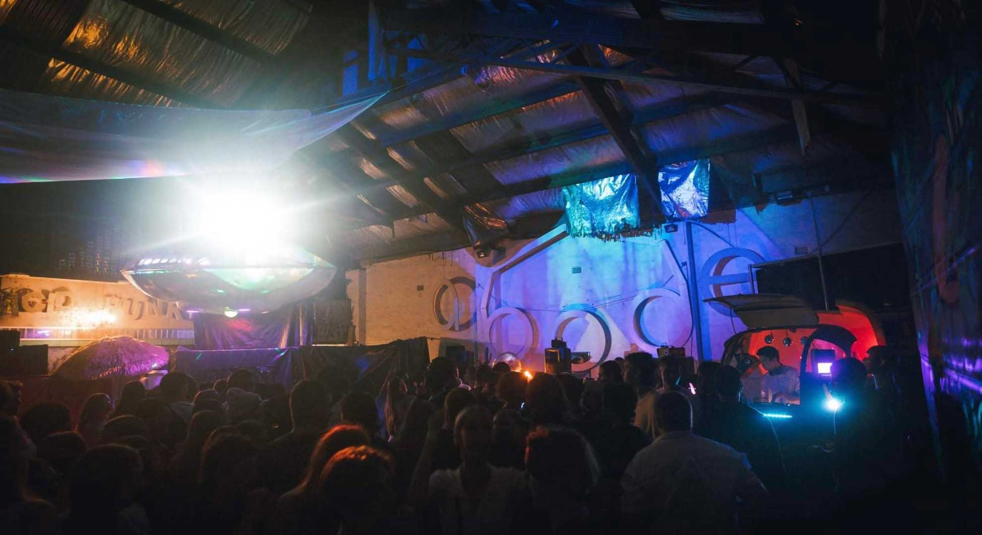 An underground warehouse with ravers watching a DJ perform in a small UFO themed stage.